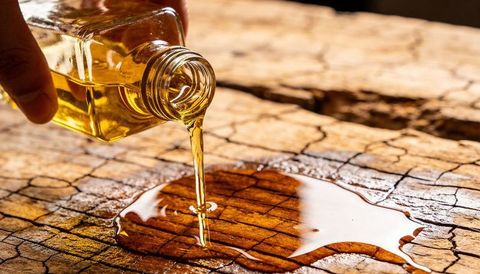 Golden oil pouring from small square glass bottle creating puddle on rustic cracked wood