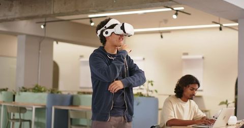African American man using VR headset as coworker typing on laptop in modern coworking
