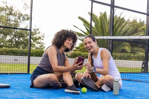 Diverse female friends sharing smartphone while resting on blue pickleball paddle court