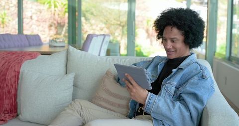 Young man relaxing and using tablet on sofa in bright modern living room near windows