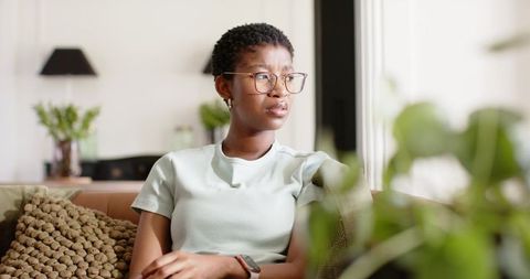 African woman sitting on sofa wearing eyeglasses and smartwatch gazing out sunlit window