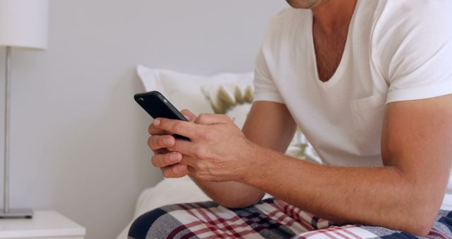 This image shows a man in casual pajamas looking at his smartphone while sitting on a bed. It can be used for lifestyle blogs, articles about morning routines, or content related to technology use at home. The image highlights themes of communication, relaxation, and staying connected.