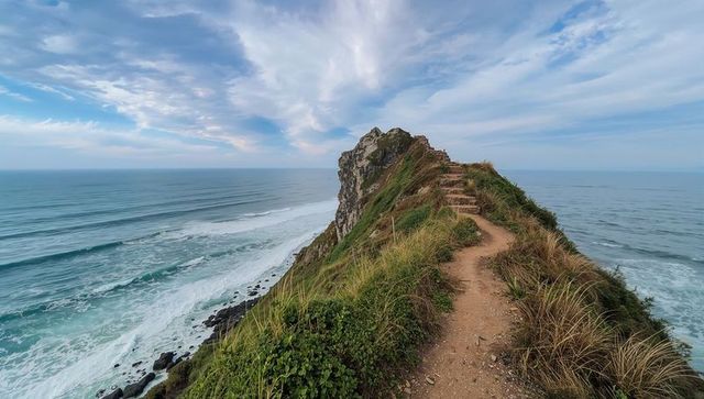 Winding coastal trail climbing narrow cliff ridge toward jagged rock outcrop over ocean