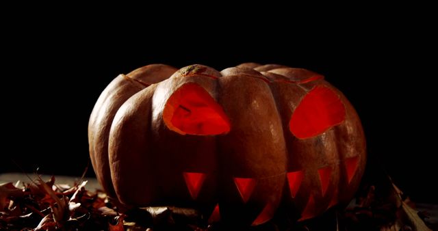 A carved pumpkin with a menacing face glows ominously in the dark, surrounded by autumn leaves. Its glowing eyes and jagged teeth create a spooky atmosphere, typical of Halloween decorations.