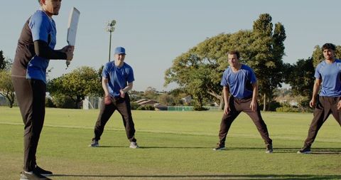 Multicultural men practicing cricket, batter poised with bat while teammates fielding on pitch
