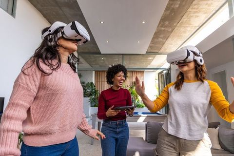 Multiracial women wearing vr headsets laughing and interacting in modern open living space