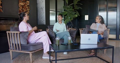 Three women discussing during casual podcast panel in modern lounge with laptop and mic