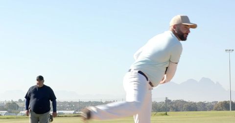 Pitcher delivering fastball with teammate walking in sunny community baseball field