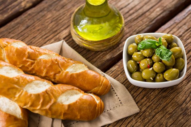 Fresh baguettes and marinated olives with a bottle of olive oil on a rustic wooden table. Ideal for use in food blogs, culinary websites, Mediterranean cuisine promotions, healthy eating articles, and gourmet recipe books.