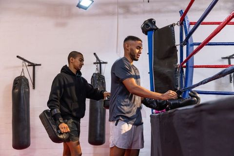 Boxing partners training in gym corner, coach holding pad, boxer adjusting black gloves