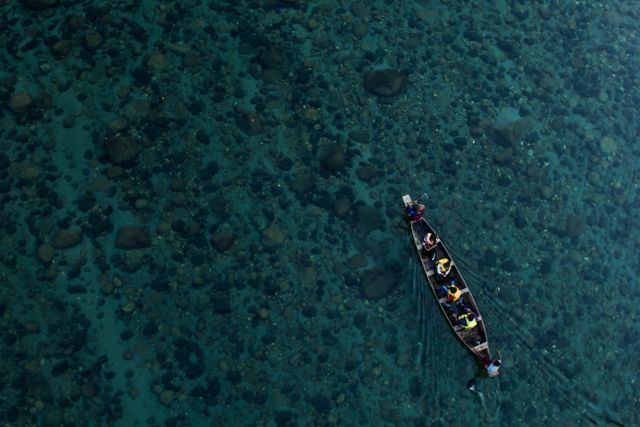 This stunning aerial view showcases people rowing a boat on a crystal clear river filled with small fishes. This scene can beautifully illustrate nature's purity through themes like outdoor adventure, scenic landscape, tranquility, and the peaceful interaction of humans with their environment. Ideal for ecotourism promotions, travel websites, and artistic natural photography collections.