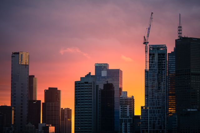 Tall buildings rise against a vibrant sunset backdrop with hues of orange and pink. A construction crane stands prominently among the skyscrapers. This image can be used for articles or projects related to urban development, architecture, city living, or evening cityscape aesthetics.