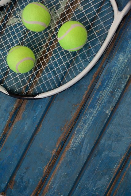 This image shows three fluorescent yellow tennis balls resting on a tennis racket placed on a blue wooden table. Ideal for use in sports-related articles, tennis equipment advertisements, or recreational activity promotions.