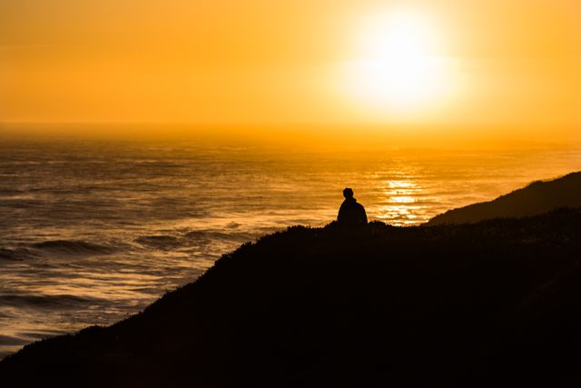 Person sitting on a clifftop overlooking the sea during sunset, highlighting a moment of peace and solitude. Warm orange and yellow hues dominate. Ideal for illustrating themes such as meditation, serenity, contemplation, and the beauty of nature's landscapes. Perfect for use in calming or inspirational content, travel marketing, and wellness promotions.