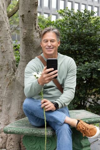 Midlife Man Sitting on Urban Bench Smiling at Smartphone While Holding White Daisies