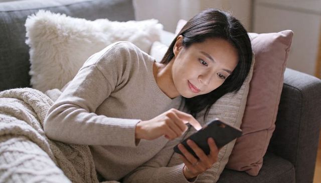 Relaxing woman scrolling smartphone on cozy sofa with knit blanket, plush cushions at home