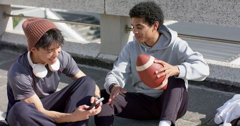 Friends sharing phone and football on rooftop, wearing hoodie and beanie