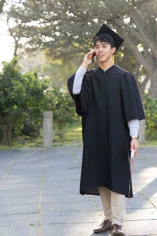 Sunlit asian graduate on campus calling on smartphone while holding diploma