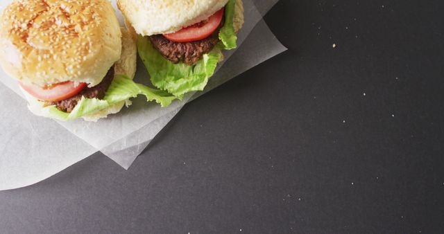 Two freshly prepared burgers placed on parchment paper with a black background, featuring lettuce, tomato, and sesame seed buns. Ideal for use in food blogs, restaurant menus, culinary articles, advertisements for lunch or meal deals, and gourmet cooking websites.