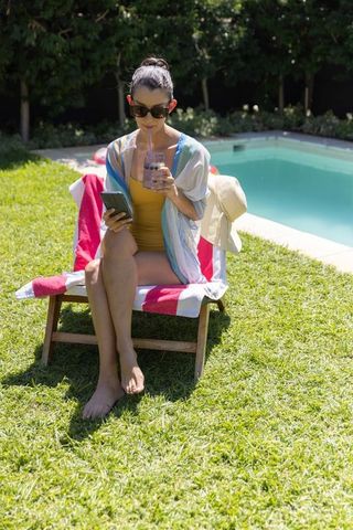 Woman relaxing by pool on lounger using phone and sipping smoothie in yellow swimsuit