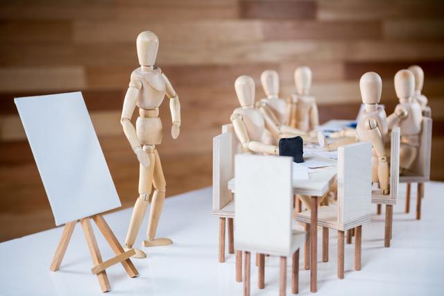 Wooden figurines are gathered around a table in a conference room, engaging in a business meeting. One figurine is standing near a blank easel, suggesting a presentation or brainstorming session. This image can be used for concepts related to teamwork, corporate training, leadership, business strategy, and creative planning.