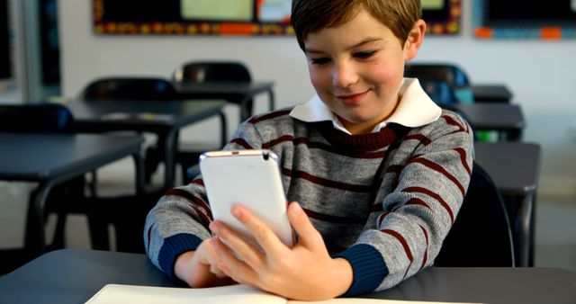 A young boy in a striped sweater is sitting at a desk in a classroom, confidently using a smartphone. The joyful expression on his face indicates enjoyment or engagement. This image is ideal for illustrating the integration of technology in education, modern classroom environments, digital literacy in children, educational apps, and the impact of mobile devices on learning.