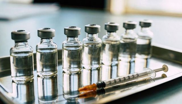 Six vaccine vials standing on stainless tray with syringes reflecting clinical sterility