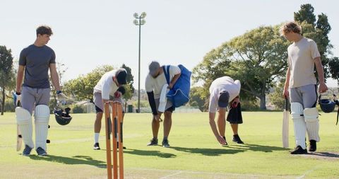 Diverse male cricket teammates preparing for training on sunlit pitch with bats, stumps