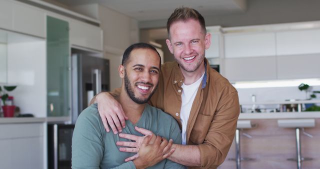 Multi ethnic gay male couple talking and embacing turning to camera and smiling. enjoying time in self isolation at home during coronavirus covid 19 pandemic.