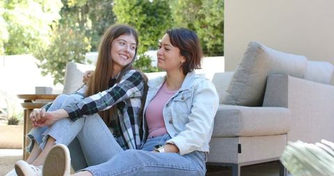 Two women relaxing on sunlit backyard patio leaning against light-gray sofa, casual denim