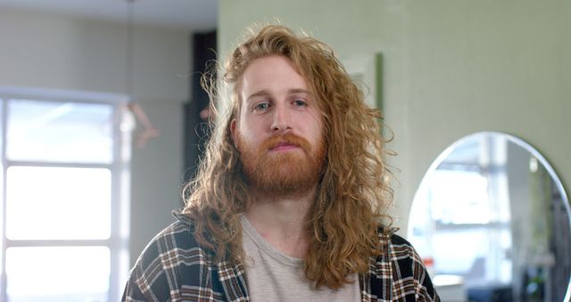 Confident man with long curly hair and beard standing indoors in casual wear. Ideal for lifestyle, fashion, and personal confidence themes. Could be used in marketing for hair care products, fashion brands, or articles about modern hairstyles. Background features soft indoor lighting and a casual home atmosphere.
