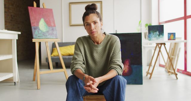 Female artist sitting in her studio with various colorful paintings displayed around the room. Art studio with canvases, showcasing creativity and modern artwork. Ideal for representing the creative process, artistic expression, and modern art environments.