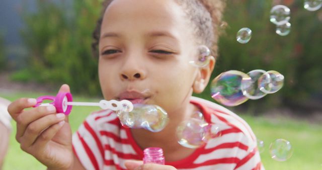 A child with curly hair wearing a striped shirt is blowing bubbles outside. The bright, sunny day and green background convey a playful, carefree scene. Ideal for usage in children's activities advertisements, summer campaign imagery, and promoting outdoor play experiences for kids.