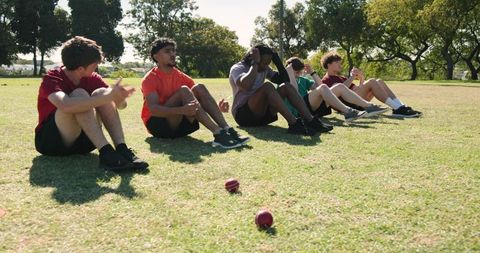 Diverse male athletes performing sit-up warm-up on park grass during outdoor team fitness training