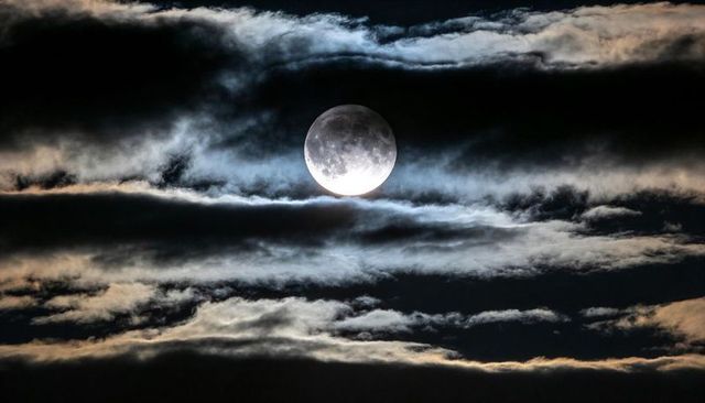 Glowing full moon revealing crater details in wide night sky with backlit layered clouds