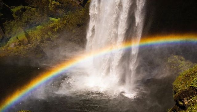 Rainbow arching across misty waterfall plunging into mossy cliff gorge, sunlit spectrum