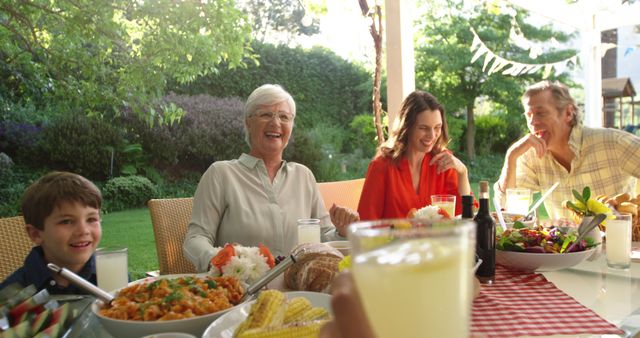 Happy multigenerational family having an outdoor meal together, enjoying each other's company in a garden setting. Perfect for advertisements and articles about family gatherings, summer activities, outdoor dining, and promoting products related to family and food enjoyment.
