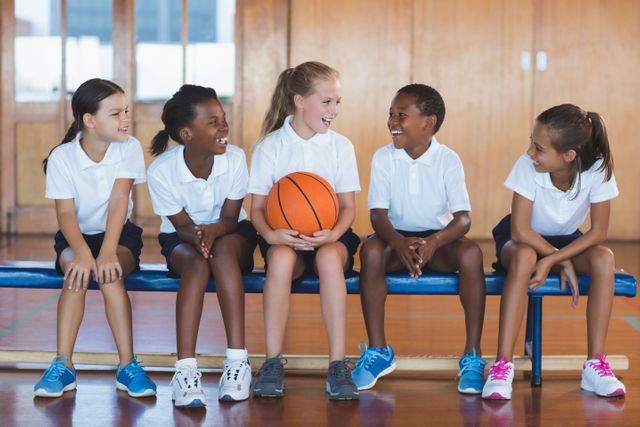 Group of diverse school kids sitting on a bench in a gym, enjoying a moment of fun with a basketball. Ideal for educational materials, sports programs, teamwork and friendship themes, and promoting physical activity among children.