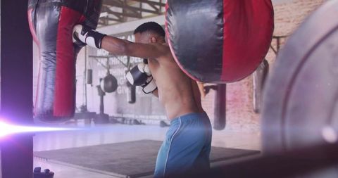 Intense shirtless boxer training with red-black heavy bag in industrial gym for stamina