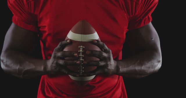 Male athlete in red football uniform holding an American football with both hands against dark background. Ideal for sports promotions, football-related advertisements, team spirit graphics, and fitness inspiration.
