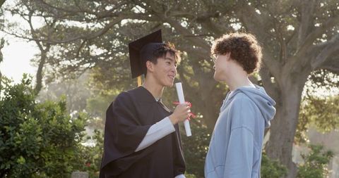 Graduate celebrating with friend on campus holding diploma wearing gown and mortarboard