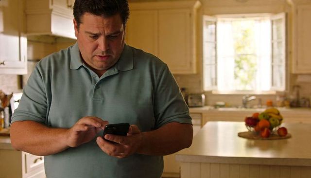 Middle-aged man tapping smartphone in sunlit kitchen with fruit bowl and focused expression