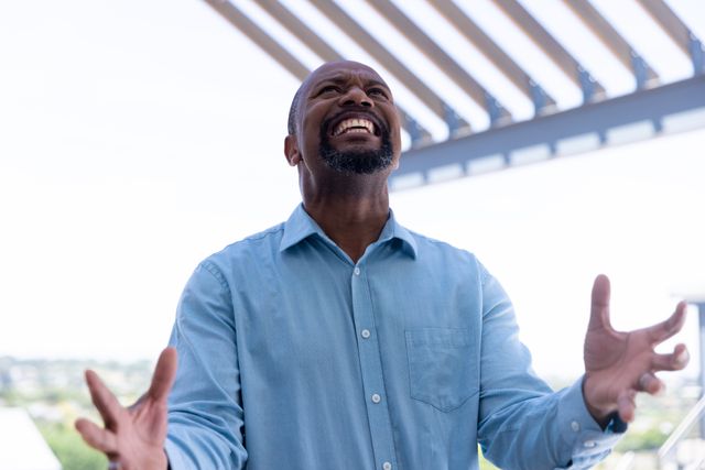 African American businessman standing outdoors with hands raised, expressing joy and success. Ideal for use in business success stories, corporate achievements, motivational content, and promotional materials for business services.
