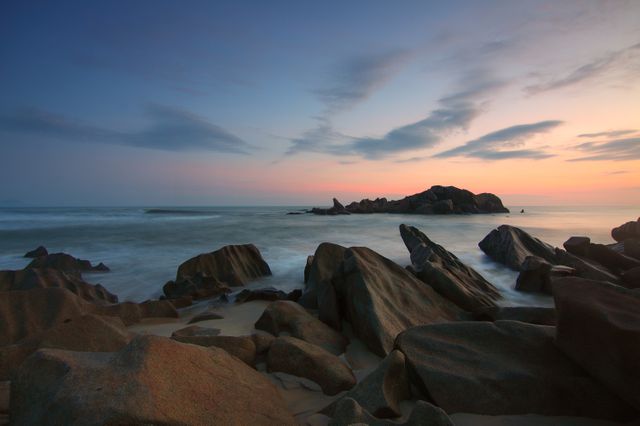 This image captures a serene coastal sunset with rugged rocks lining the shore and calm ocean waters. Use this for nature-themed projects, relaxing backgrounds, or travel promotions highlighting coastal regions.