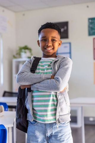 Smiling african american preteen standing with arms crossed in classroom carrying backpack