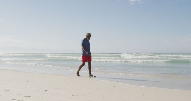 Senior man walking alone on sandy beach, wearing red shorts and blue shirt. Calm waves in background and clear blue sky. Ideal for themes like retirement, relaxation, summer holidays, and peaceful coastal scenes.