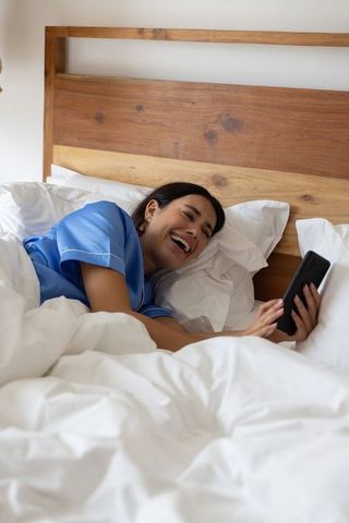 Smiling woman lounging in bed using smartphone, cozy white bedding, wooden headboard