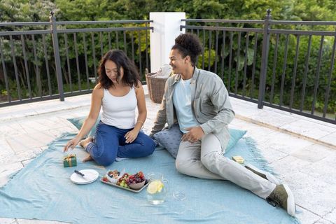 Rooftop picnic with diverse couple sharing fruit, gift and lemonade on light-blue blanket