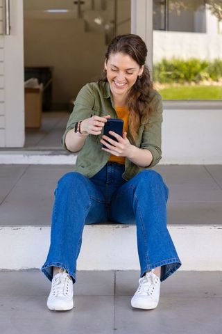 Smiling woman sitting on stoop scrolling smartphone while wearing smartwatch and casual jeans