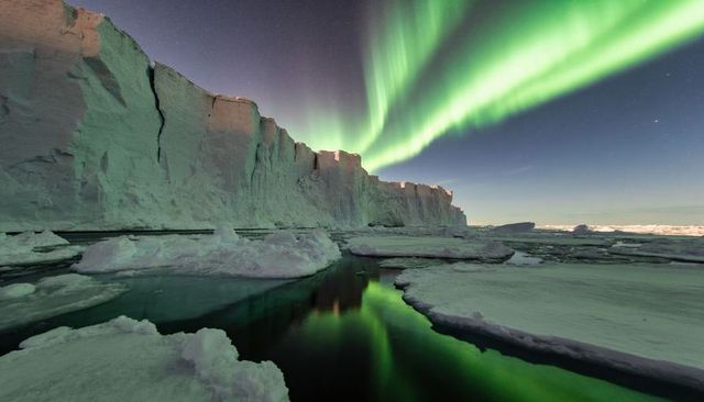Green aurora glowing over towering polar ice cliff reflecting on dark arctic water